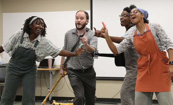 A photo of 4 musicians singing in a classroom. A man holds a banjo while a woman holds a guitar.
